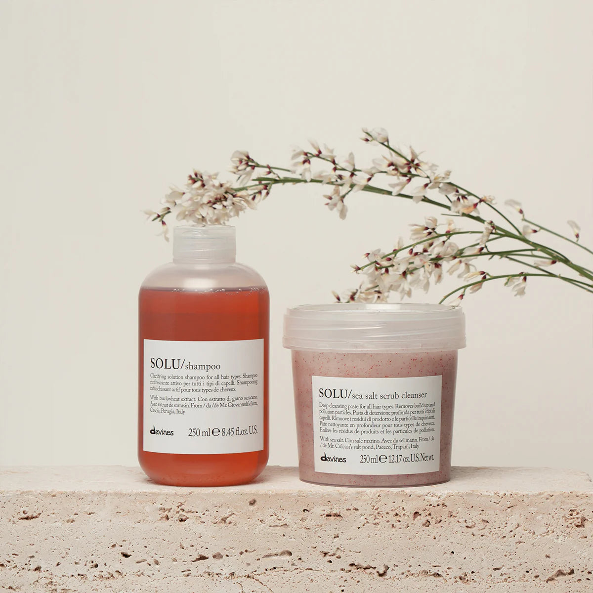 Davines Solu shampoo and Davines Solu scrub cleanser container with flowers on a neutral background