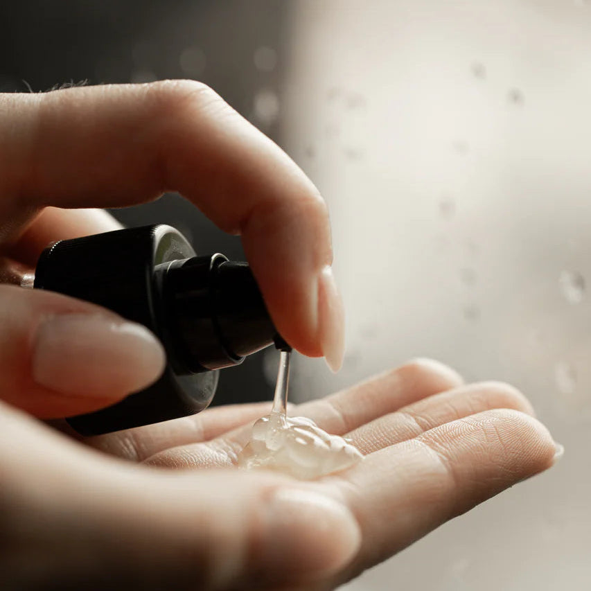Hand holding a black spray bottle over another hand with a light-coloured Smoothing Fluid gel being sprayed.