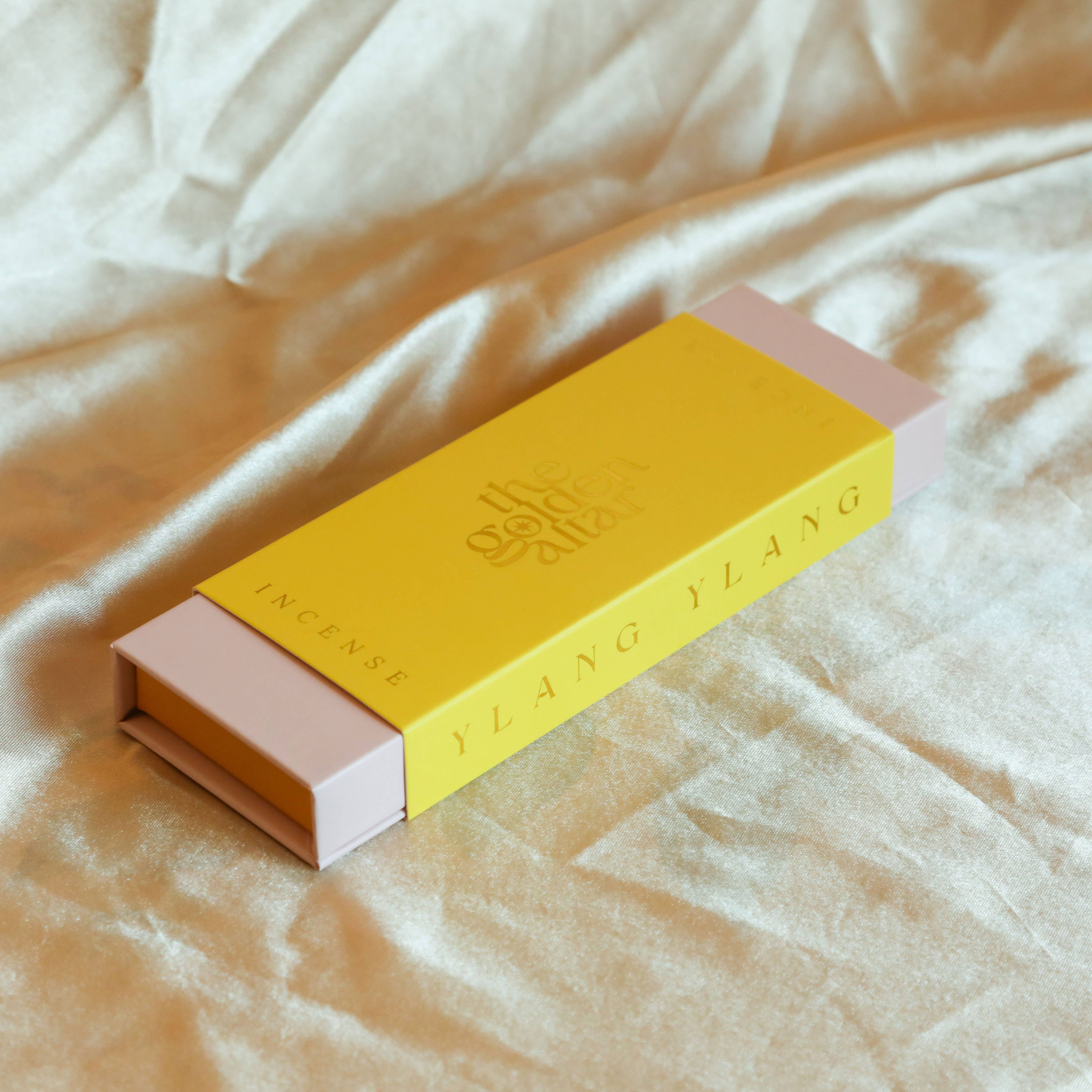 Yellow and pink incense box labeled The Golden Altar with scent 'Ylang Ylang' on a beige fabric background