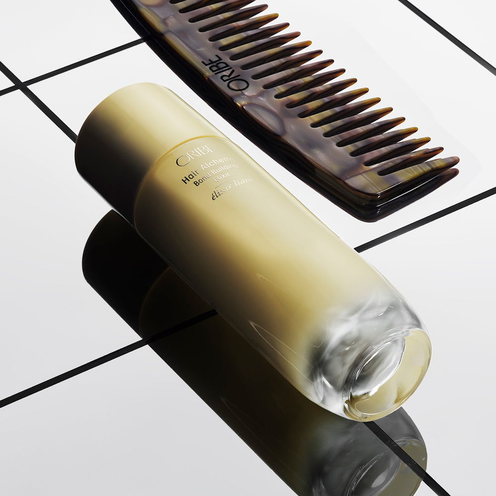Oribe Hair Alchemy Bond Building Elixir glass bottle lying flat beside an Oribe comb on a tiled surface