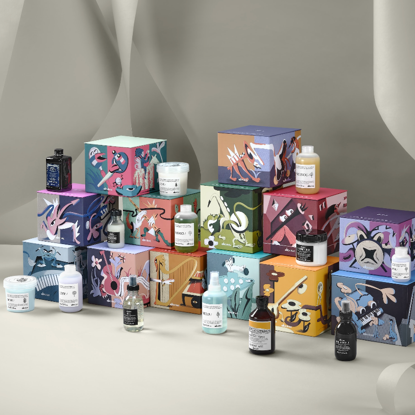 A curated collection of Davines Gift products arranged in stacked boxes on a draped beige studio set. The products include bottles, jars, and containers with illustrated packaging in shades of blue, pink, purple, green, orange, and black, alongside white jars and small black bottles.