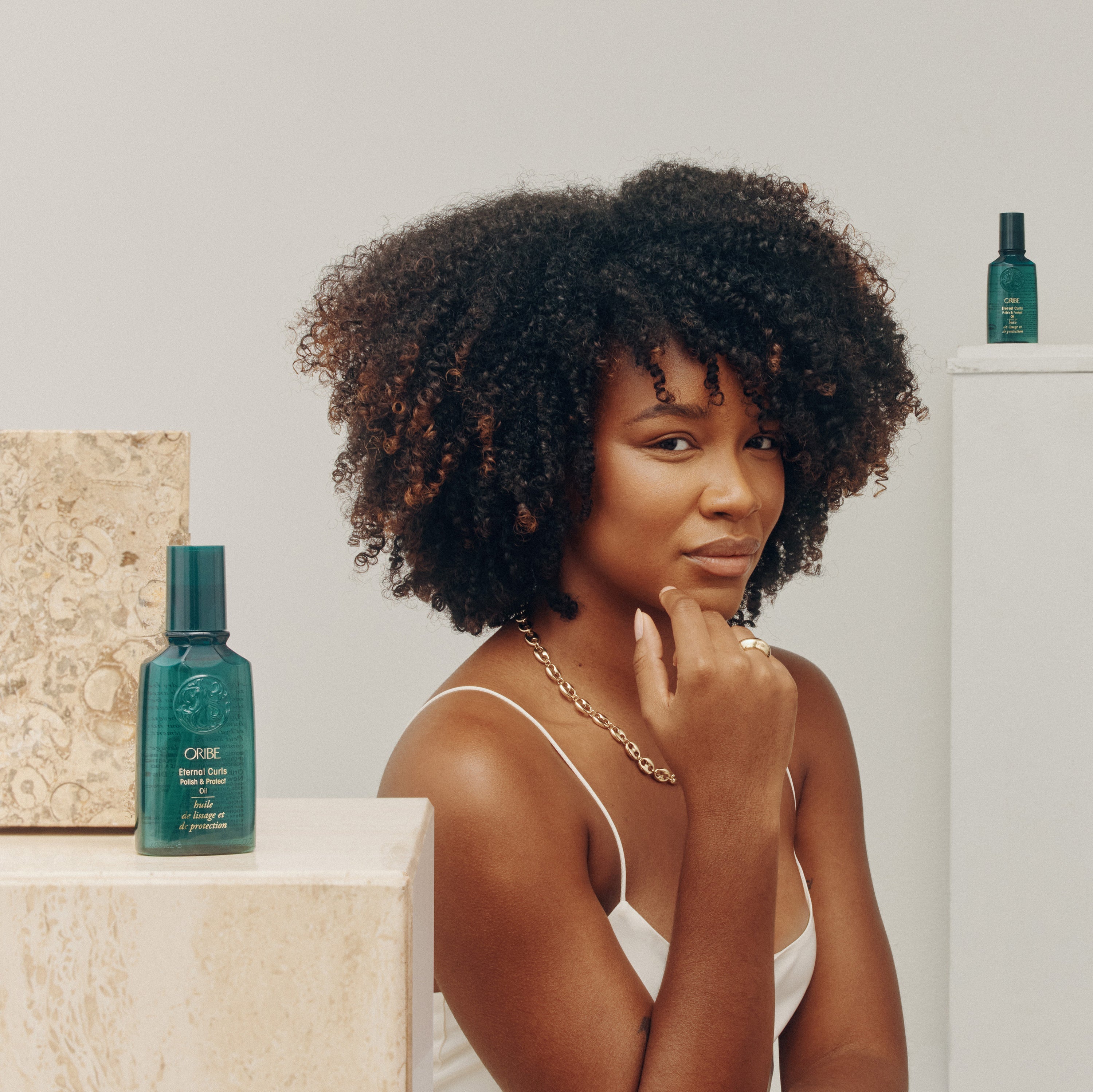 Woman with curly hair sitting next to a bottle of Oribe Eternal Curls Polish and Protect Oil on a white background
