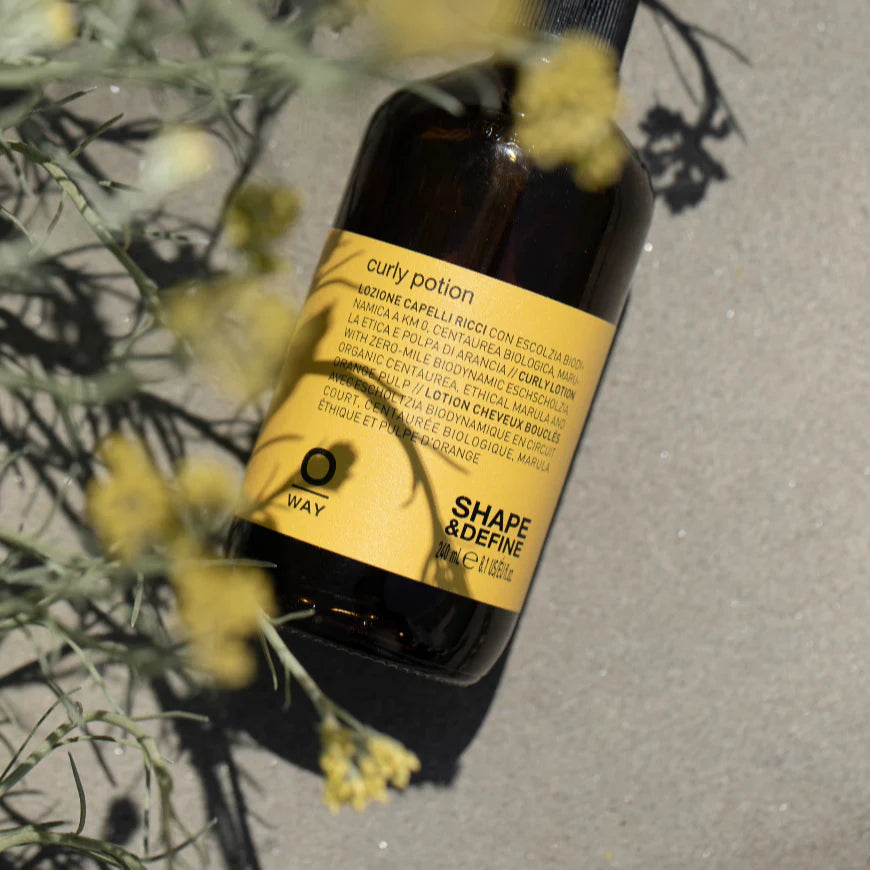 Bottle of 'Oway Curly Potion' with a yellow label on a gray surface with plants.